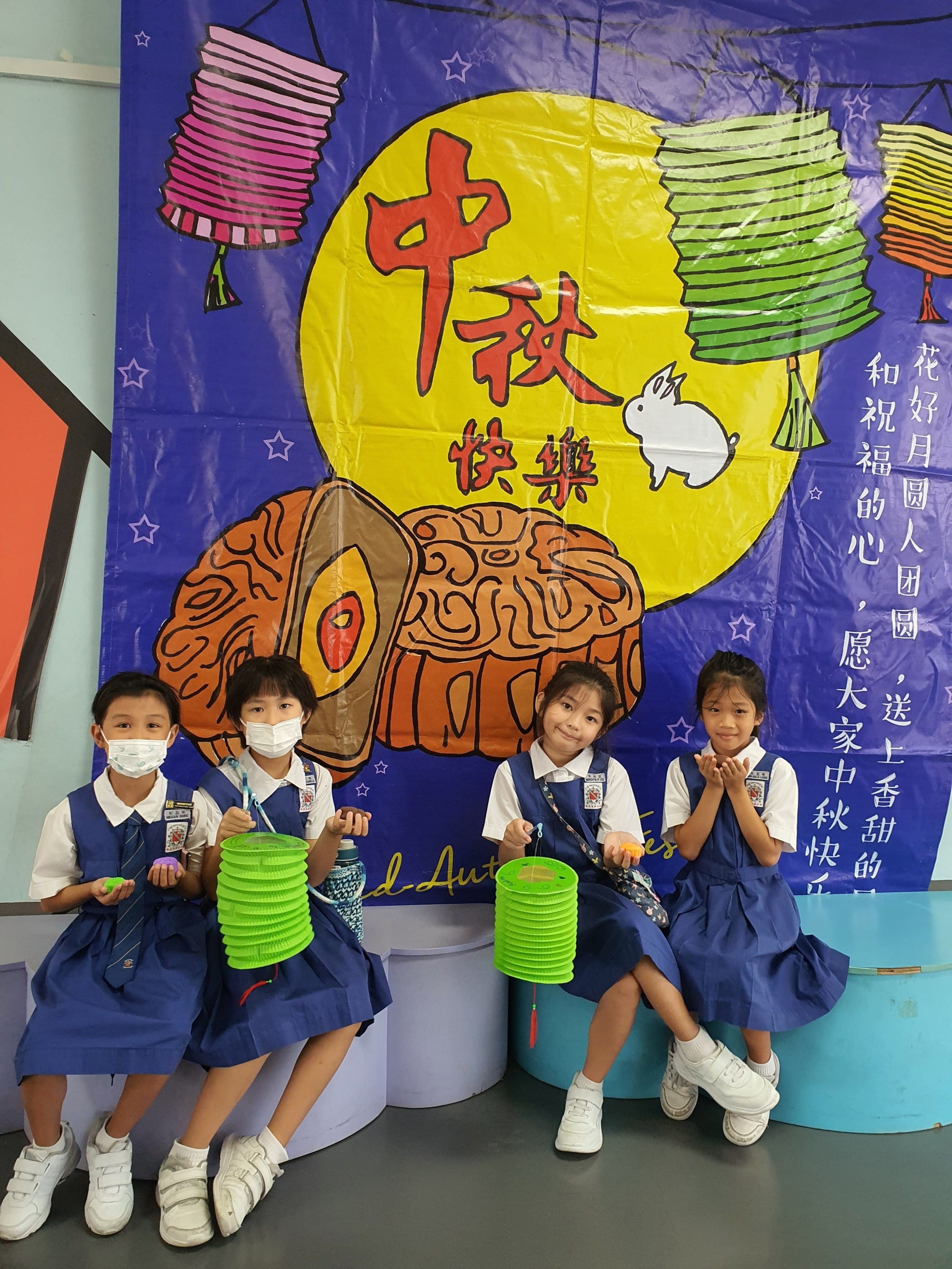 Four young children in blue uniforms hold green lanterns in front of a colorful Mid-Autumn Festival banner with Chinese text.